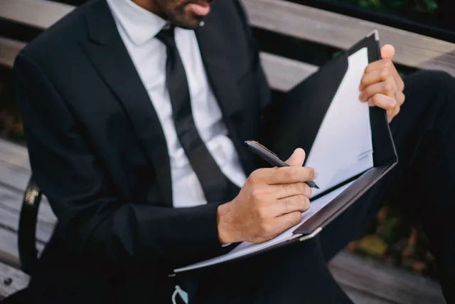 A businessman in a suit writing on a clipboard while sitting on a wooden bench outdoors. Mobile notary software options, reviewed and ranked