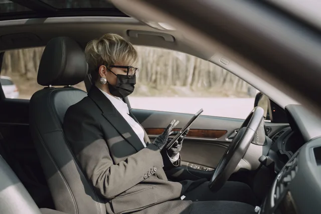 Side view of concentrated adult woman in formal clothes and protective mask and gloves sitting on driver seat of contemporary car and using tablet. Mobile notary startup: the eight steps, in order