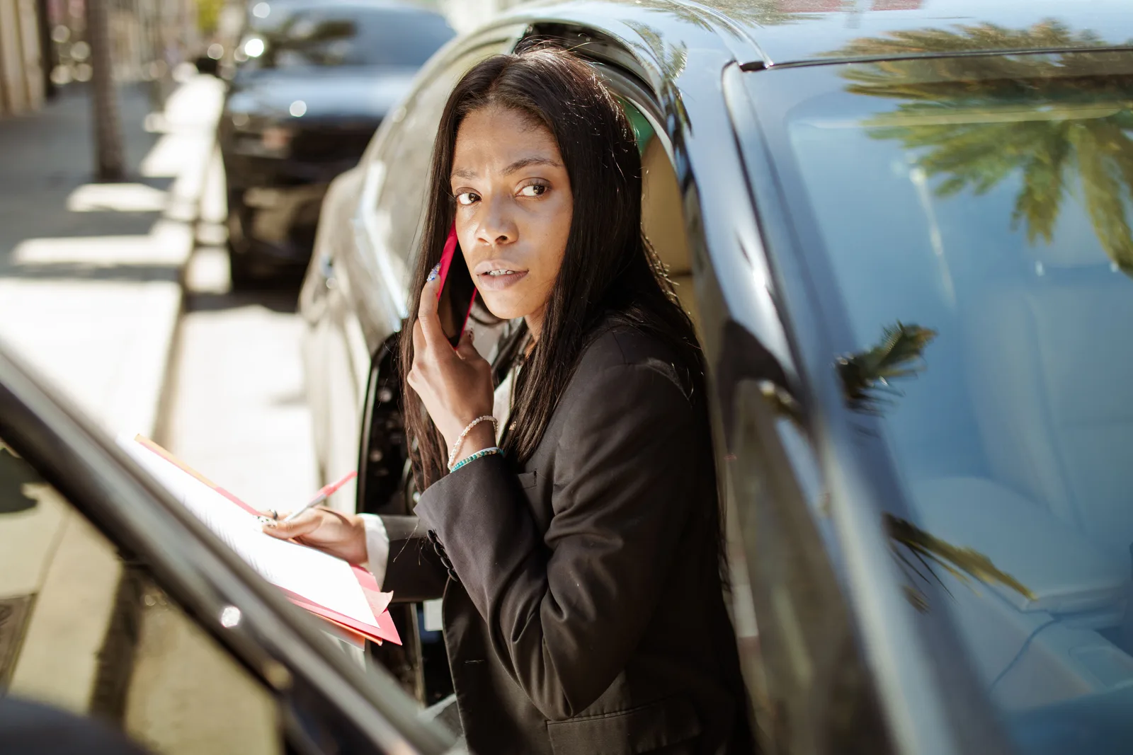 Confident businesswoman in corporate attire multitasking with a phone call and notes in a car. Measuring profitable mobile notary services